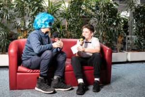 Ein Alien auf der Interviewcouch. Zwei Jungs sitzen auf einem roten Ledersofa. Einer von ihnen ist als Alien verkleidet und wird von dem anderen interviewt.